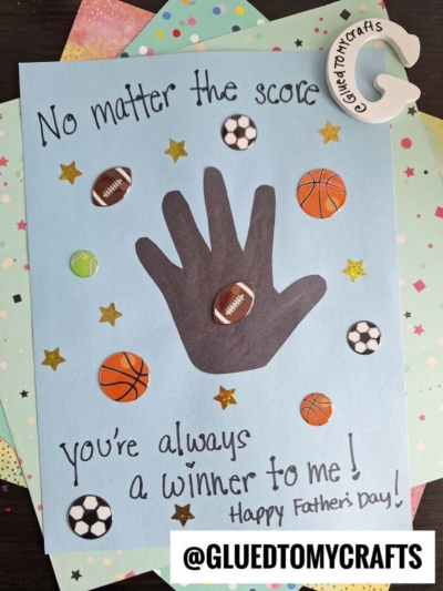 A handmade HANDPRINT FATHER'S DAY card features a black handprint, sports ball stickers, gold stars, and the message: “No matter the score, you’re always a winner to me! Happy Father’s Day!” Craft materials are in the background.