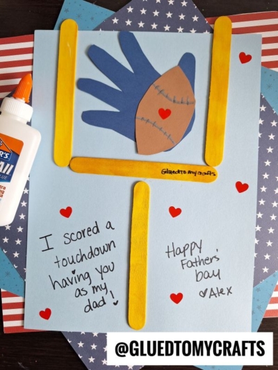 A handmade HANDPRINT FATHER'S DAY card features a football goalpost made from yellow craft sticks, a handprint football, red hearts, and handwritten messages on blue paper. A bottle of glue and a patriotic background are visible.