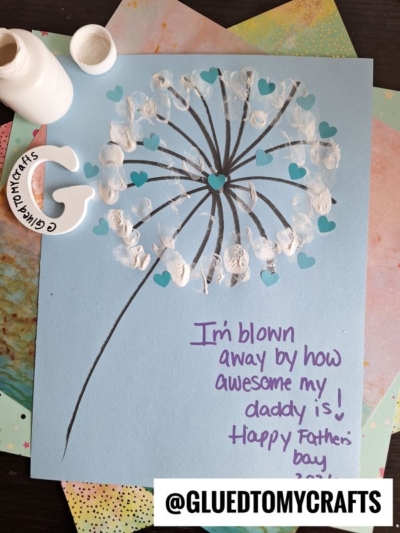 A handmade HANDPRINT FATHER'S DAY card featuring a dandelion made from white fingerprints and teal heart confetti on blue paper. It reads, I'm blown away by how awesome my daddy is! Happy Father's Day. Craft supplies surround the card.