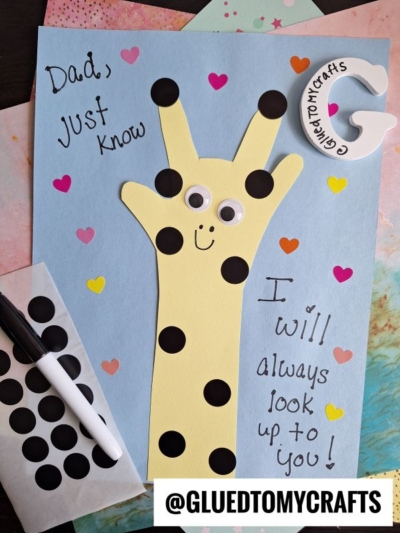 A handmade HANDPRINT FATHER'S DAY card featuring a yellow giraffe crafted from a traced hand with googly eyes and black spots, surrounded by colorful hearts. The card reads, Dad, just know I will always look up to you!.