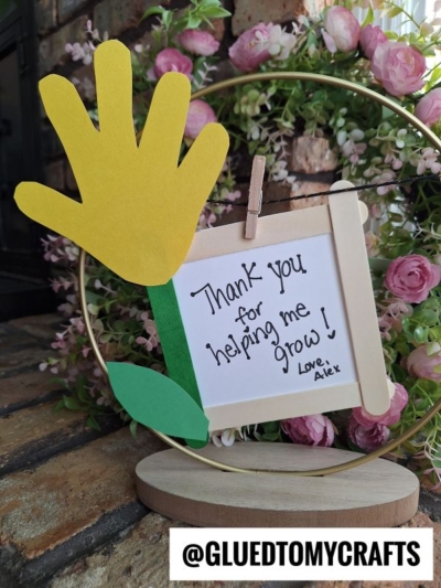 A handmade HANDPRINT FATHER'S DAY craft featuring a yellow paper handprint, green stem and leaf, and a "Thank you for helping me grow! Love, Alex" sign, displayed among pink flowers with @GLUEDTOMYCRAFTS at the bottom.