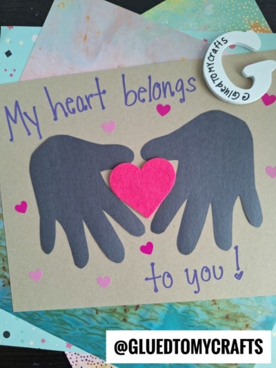 A handmade HANDPRINT FATHER'S DAY card with black paper cutouts of two hands holding a red heart, surrounded by pink hearts and the words “My heart belongs to you!” on brown. The @GLUEDTOMYCRAFTS label is at the bottom.