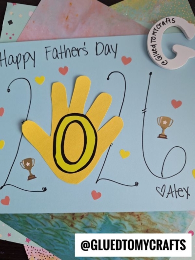 A handmade HANDPRINT FATHER'S DAY card reads Happy Fathers Day 2026, with the 0 shaped from a yellow handprint. Decorated with hearts and trophies, it's signed Alex. A @GluedToMyCrafts sticker is placed at the bottom.