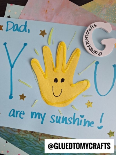 A handmade HANDPRINT FATHER'S DAY card with "Dad, you are my sunshine!" in blue and green, featuring a yellow handprint smiley, gold star stickers, and a pink paper G. @GLUETOMYCRAFTS watermark is at the bottom.