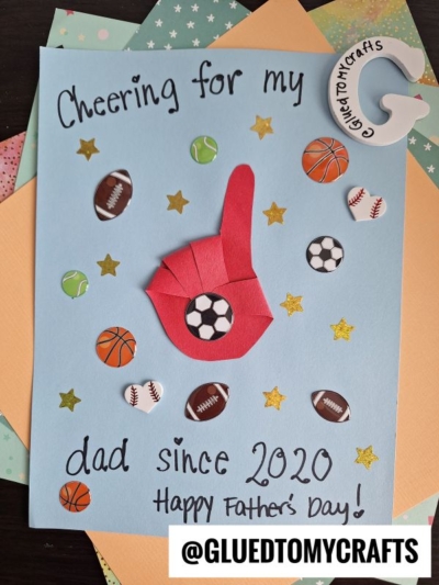 A handmade HANDPRINT FATHER'S DAY card with "Cheering for my dad since 2020" on blue paper, decorated with sports ball stickers, gold stars, a red foam finger, and a craft badge. Makers handle: @GLUEDTOMYCRAFTS.