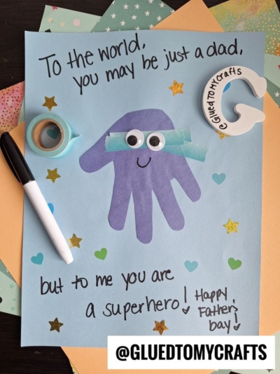 A handmade HANDPRINT FATHER'S DAY card features a purple handprint with googly eyes and a blue paper mask, resembling a superhero. The card says, To the world, you may be just a dad, but to me you are a superhero! Happy Fathers Day!.