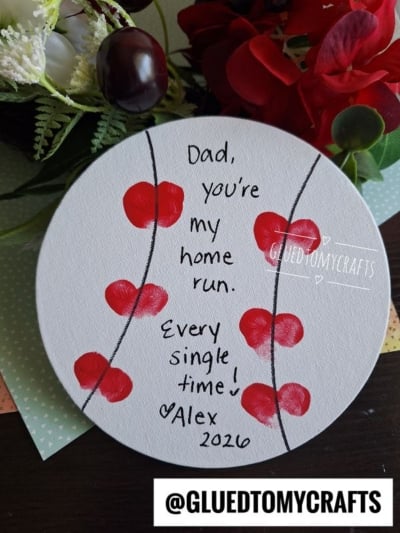 A round canvas painted like a baseball with red thumbprint hearts—perfect for THUMBPRINT CRAFTS FOR KIDS. Text reads: Dad, you’re my home run. Every single time! ♥ Alex 2020. Flowers and craft supplies are in the background.