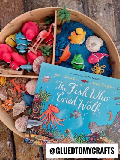 A round tray with toy sea animals, blue sand, and shells sits on a brick surface beside the book The Fish Who Cried Wolf by Julia Donaldson—perfect for story time sensory bin ideas. The @GLUEDTOMYCRAFTS handle appears at the bottom.
