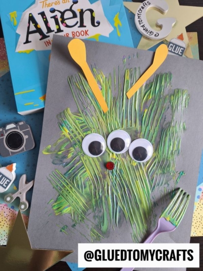 A green and yellow fork painted alien face made with paint, googly eyes, and craft sticks on gray paper. Surrounded by craft supplies, a book titled “There’s an Alien in Your Book,” and a tag reading @GLUEDTOMYCRAFTS.