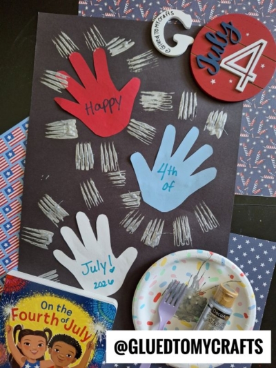 A July 4th craft with red, white, and blue paper handprints on black paper, chalk fireworks, a book, paint supplies, and patriotic decorations—perfect for creating 4th of July keepsakes. The handle @GLUEDTOMYCRAFTS is at the bottom.