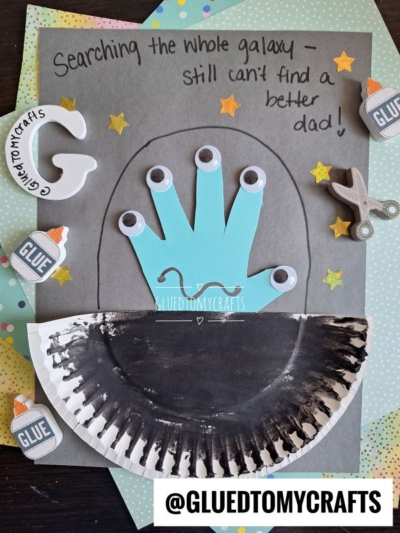 A handmade HANDPRINT FATHER'S DAY card features a blue handprint alien with googly eyes, a black half-circle spaceship, star stickers, and the message: Searching the whole galaxy – still can’t find a better dad!.