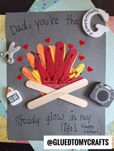 A HANDPRINT FATHER'S DAY craft with paper flames and popsicle stick logs on black paper. Surrounded by stickers of glue, scissors, camera, and a craft tag. Text reads, “Dad, you’re the steady glow in my life! Happy Father’s Day.”.