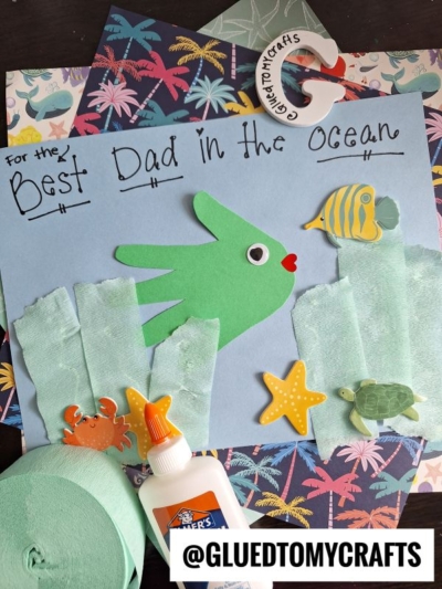 A handmade HANDPRINT FATHER'S DAY card featuring a green handprint fish with yellow fins and a googly eye, surrounded by sea creatures, tissue paper seaweed, and the message “For the Best Dad in the Ocean.” Craft supplies add a creative touch.