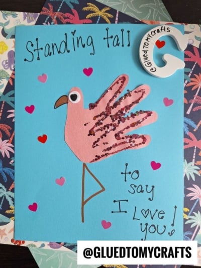 A handmade flamingo card with glitter, a googly eye, and heart stickers on blue paper—perfect for a HANDPRINT FATHER'S DAY surprise. The text says, Standing tall to say I love you! and a tag reads @GLUEDTOMYCRAFTS.