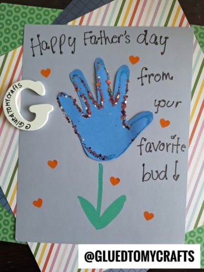 A handmade HANDPRINT FATHER'S DAY card featuring a blue handprint outlined in glitter, orange hearts, and a green paper stem. The striped background reads “Happy Father’s Day from your favorite bud!”.