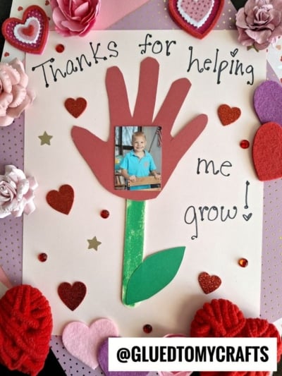 A handmade HANDPRINT FATHER'S DAY card featuring a child’s photo centered on a red handprint flower with a green stem and leaves. The card reads “Thanks for helping me grow!” and is decorated with hearts, flowers, and sparkles.