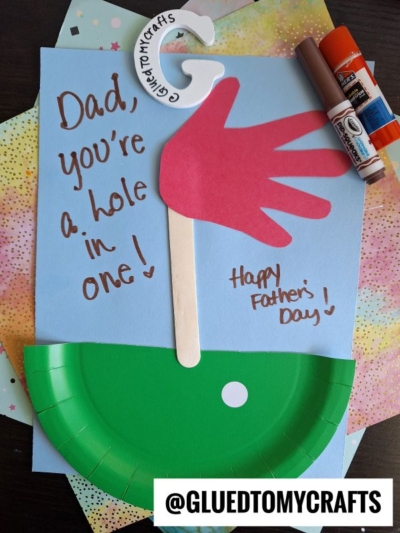 A HANDPRINT FATHER'S DAY card with a golf theme features a red handprint as a golf club, a green paper plate for the hole, and the message "Dad, you’re a hole in one!" Markers and glue add creative touches nearby.