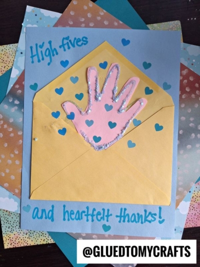 A handmade HANDPRINT FATHER'S DAY card with "High fives and heartfelt thanks!" in blue, a pink handprint cutout inside a yellow envelope, and blue hearts. The @GLUEDTOMYCRAFTS handle is at the bottom.
