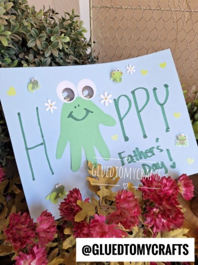 A handmade HANDPRINT FATHER'S DAY card features a green handprint frog with googly eyes, flower stickers, and “HAPPY Fathers Day!” among small frog and heart decorations. Pink flowers and greenery are in the foreground.