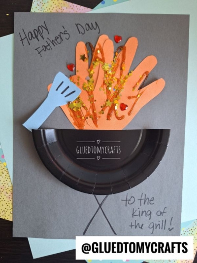 Handmade HANDPRINT FATHER'S DAY card featuring paper cutouts of handprints as flames in a black paper plate grill, with a blue paper spatula, glitter decorations, and text: “Happy Father’s Day” and “to the king of the grill!”.