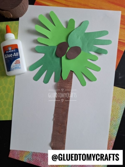A handprint palm tree craft made from green handprint cutouts for leaves, brown paper for the trunk, and two brown ovals for coconuts, with a bottle of glue on the side and @GLUETOMYCRAFTS at the bottom.