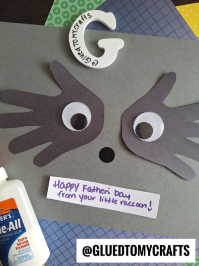 A HANDPRINT FATHER'S DAY raccoon craft features black handprint cutouts as the raccoon's face, large googly eyes, a white paper G, and a note reading Happy Father's Day from your little raccoon! A glue bottle is nearby.