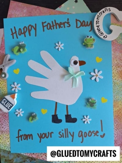 A handmade HANDPRINT FATHER'S DAY card features a goose crafted from a white handprint cutout on blue paper, decorated with a bow, beak, and boots, surrounded by frog stickers, hearts, and the message “Happy Father’s Day from your silly goose!”.