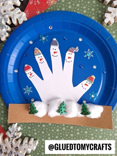A blue paper plate creates a charming handprint snowman snow globe, with each white finger styled as a snowman in hats and scarves. Cotton balls and mini Christmas trees line the base, while snowflakes and gems accent the frosty scene.