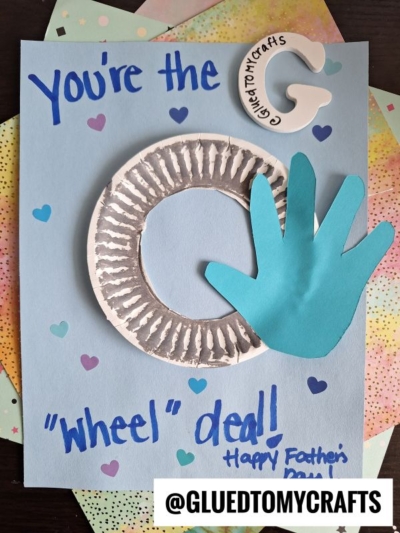 A handmade HANDPRINT FATHER'S DAY card features the message You’re the G ‘wheel’ deal! Happy Father’s Day, with a paper plate wheel, a blue cut-out hand, and colorful heart stickers on a blue background.
