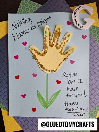 A HANDPRINT FATHER'S DAY card with a yellow handprint flower outlined in gold glitter. It reads, Nothing blooms as bright as the love I have for you! Happy Father’s Day! 2020. Hearts and a green stem complete this heartfelt keepsake.