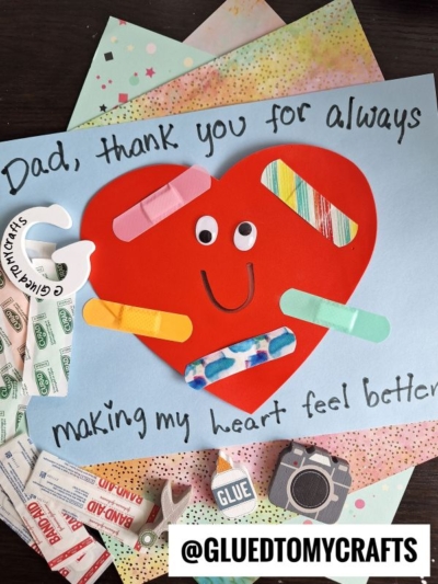 A HANDPRINT FATHER'S DAY card reads Dad, thank you for always making my heart feel better with a big red heart, googly eyes, smile, and colorful bandages. Craft supplies, glue, and stickers are scattered around.