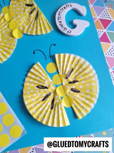 A yellow butterfly made from folded polka-dot cupcake liners and paper circles is glued to blue paper. Black lines are drawn for antennae and details. The @GLUEDTOMYCRAFTS handle appears at the bottom.