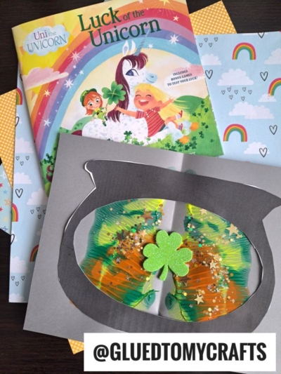 A colorful children’s book titled Luck of the Unicorn sits above a St. Patrick’s Day-themed craft featuring a paper pot of gold, green glitter, and a foam shamrock; PAINT SPLAT CRAFTS and patterned papers brighten the background.
