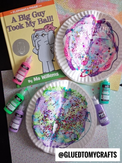 Children’s book “A Big Guy Took My Ball!” is shown with two painted paper plates, playful paint splat designs, glitter, and bottles of colorful paint, set on a gray surface. Art supplies are scattered around, and the @GLUEDTOMYCRAFTS watermark is visible.