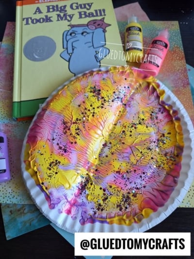 A paper plate painted with pink, yellow, and white swirls sits near a children’s book titled “A Big Guy Took My Ball!” and bottles of craft paint. The plate is decorated with splatters and marbled patterns.