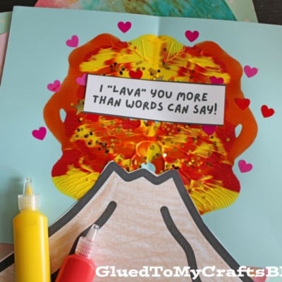 A handmade volcano craft with red, yellow, and orange paint, decorated with small heart stickers and a sign reading I LAVA YOU MORE THAN WORDS CAN SAY! Bottles of red and yellow paint sit beside the craft.