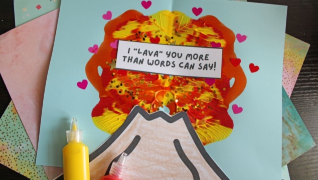 A handmade volcano craft with red, yellow, and orange paint, decorated with small heart stickers and a sign reading I LAVA YOU MORE THAN WORDS CAN SAY! Bottles of red and yellow paint sit beside the craft.