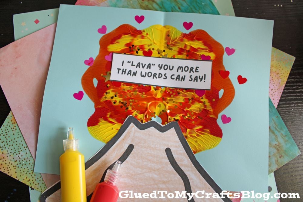 A handmade volcano craft with red, yellow, and orange paint, decorated with small heart stickers and a sign reading I LAVA YOU MORE THAN WORDS CAN SAY! Bottles of red and yellow paint sit beside the craft.