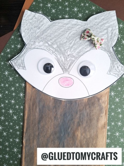 A handmade lunch sack wolf puppet with googly eyes, a pink nose, and a small floral bow sits on a green star-patterned background. The handle @GLUEDTOMYCRAFTS is displayed at the bottom.