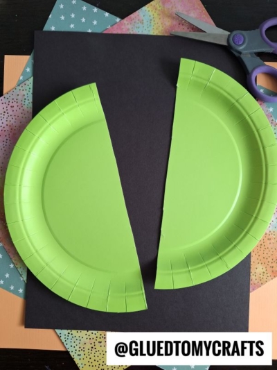 A green paper plate cut in half is placed on black paper, surrounded by colorful sheets and scissors. The Instagram handle @GLUEDTOMYCRAFTS appears at the bottom.