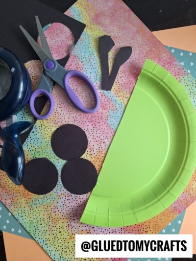 Colorful craft supplies, including patterned paper, green paper plate, black paper circles, scissors, and a hole punch, arranged on a table. The handle @GLUEDTOMYCRAFTS is visible in the bottom right corner.