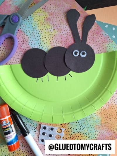 A green paper plate decorated with three black paper circles, two paper ears, and googly eyes to make a bunny, surrounded by craft supplies like scissors, glue, and more googly eyes. @GLUEDTOMYCRAFTS is at the bottom.