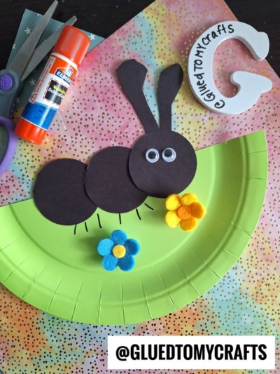 A craft project of a bunny made from black paper circles, googly eyes, and yellow and blue felt flowers on a green paper plate. Scissors, glue, and other craft materials are nearby. @GluedToMyCrafts is written at the bottom.