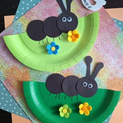 Two paper plate crafts shaped like ants, made with green plates, black paper circles, felt flowers, and googly eyes, are displayed on patterned paper backgrounds. @GLUEDTOMYCRAFTS is written at the bottom.