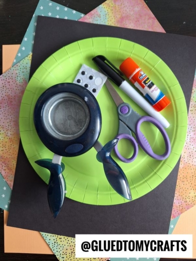 A green paper plate holds scissors, a glue stick, a black marker, a hole punch, and googly eyes. The plate is on top of colorful and black sheets of paper. @GLUEDTOMYCRAFTS is written at the bottom.