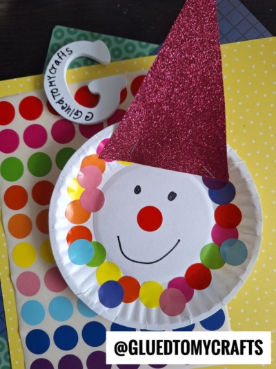 A paper plate clown craft with a red glittery hat, multicolored dot stickers for hair, and a drawn face sits on colorful polka dot paper. Perfect for coding label crafts, @GLUEDTOMYCRAFTS is written at the bottom.