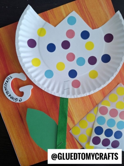 A vibrant paper plate tulip decorated with colorful dot stickers and a green paper stem and leaf, displayed on a wooden surface with extra dot stickers and a craft logo nearby.