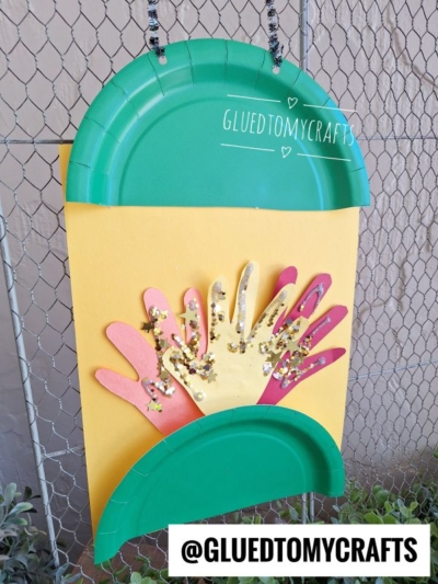 A lantern craft made from green paper plates and yellow paper, featuring cut-out hand shapes as the flames decorated with glitter, hanging on a fence. The text “GLUEDTOMYCRAFTS” is on the yellow paper and bottom corner.