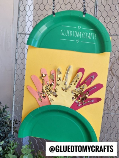 A yellow card with colorful paper hand cutouts and gold glitter, framed by green paper plates, hangs on a fence. The top plate says GLUEDTOMYCRAFTS and the handle @GLUEDTOMYCRAFTS is at the bottom.