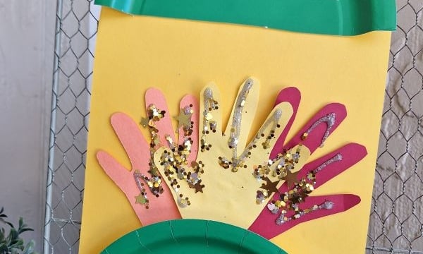 A yellow card with colorful paper hand cutouts and gold glitter, framed by green paper plates, hangs on a fence. The top plate says GLUEDTOMYCRAFTS and the handle @GLUEDTOMYCRAFTS is at the bottom.